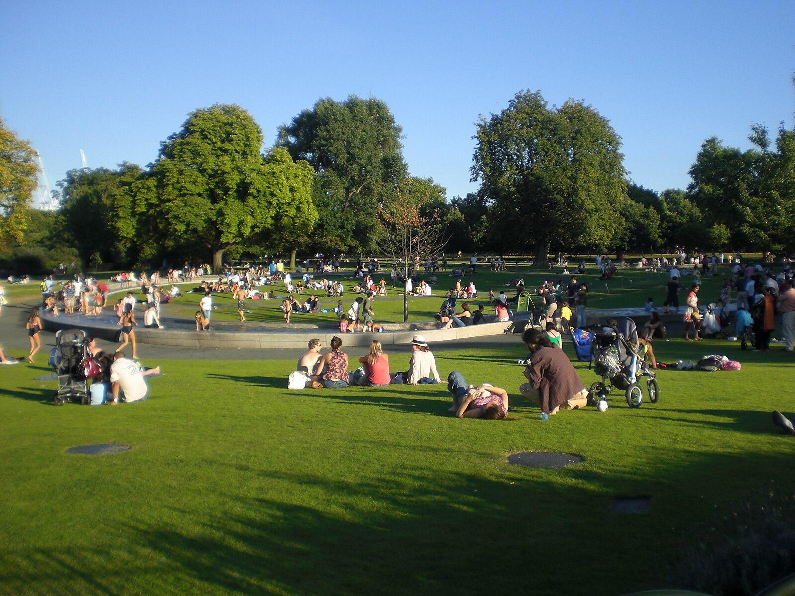 People of all ages gather on the green surrounding Diana Memorial Fountain. Some sit in groups and others enter the fountain water in swimsuits. Image Credit: Wikimedia Commons